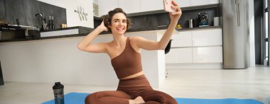 Yaşam tarzı ve egzersiz. Modern fitness kızı yoga minderinde selfie çekiyor, egzersizlerini evde yapıyor ve akıllı telefondan fotoğraf çekiyor, canlı video, telefonda video kaydediyor..