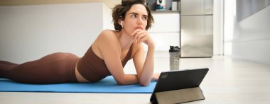Düşünceli yüzlü fitness kızı portresi, evdeki kauçuk yoga minderine uzanmış, tablet üzerinde spor videoları izleyip, düşünüyor..