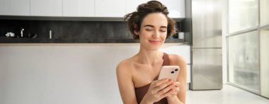 Spor ve kadın yaşam tarzı. Genç bir kadın evde egzersiz yapıyor, mutfakta akıllı telefonuyla yoga minderinde oturuyor, aktif kıyafet giyiyor, ev içi egzersizler için cep telefonu uygulaması kullanıyor..