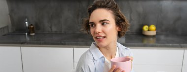 Evde kahve içen mutlu esmer kadın. Pembe bardaklı kız, mutfağında oturuyor ve hafta sonunun keyfini çıkarıyor..