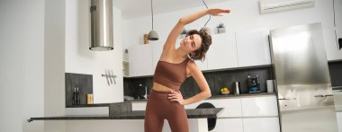 Ev ödevi konsepti. Spor elbiseli genç bir kadın, esneme egzersizleri yapıyor, mutfakta antrenman yapıyor, kolunu bir tarafa çekiyor, vücuduna odaklanıyor. Boşluğu kopyala