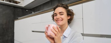 Şefkat ve rahatlama. Genç bir kadın kahve içiyor ve gülümsüyor, bir fincan çayla rahatlıyor, mutfakta yerde oturuyor..