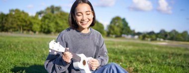 Kaygısız Asyalı kız parkta ukulele çalıp şarkı söylüyor, çimlerin üzerinde oturuyor, müzisyen boş zamanlarında dışarıda dinleniyor..