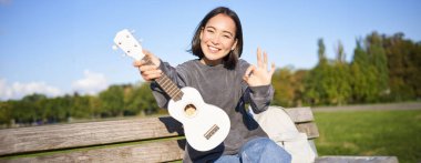 Sevimli gülümseyen kız tamam işareti ve yeni ukulelesi, parkta bankta oturuyor, müzik aleti tavsiye ediyor..