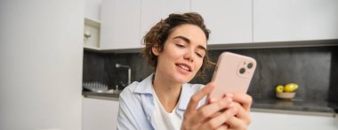 Cep telefonu tutan ve gülümseyen modern esmer kadının portresi. Kız evde akıllı telefonuyla mutfakta oturuyor, teslimat için uygulama kullanıyor, online alışveriş yapıyor..