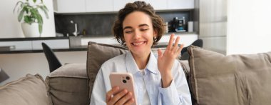 Arkadaş canlısı bir kız cep telefonuyla konuşur, arkadaşıyla görüntülü sohbetler yapar, akıllı telefon kamerasına el sallar, evdeki kanepede oturur..