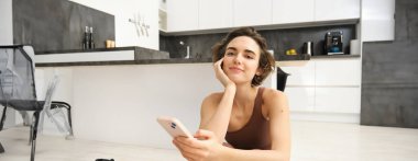 Teknoloji ve spor konsepti. Jimnastikçi kız akıllı telefonuna bakıyor, spor kıyafetleriyle evde oturuyor, cep telefonuyla egzersiz videoları izliyor..
