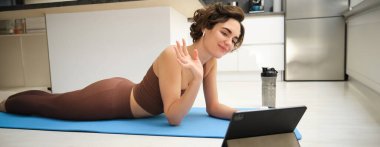 Spor ve yaşam tarzı. Genç fitness kadını, beden eğitimi hocası tablette el sallıyor, çevrimiçi video sınıfına katılıyor, yoga ve farkındalık egzersizi yapıyor, evde antrenman yapıyor..