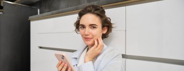 Cep telefonlu bir kızın portresi, evde oturuyor, uygulamadan sipariş veriyor. Akıllı telefonlu genç bir kadın evden online alışveriş yapıyor, web sitelerine bakıyor..