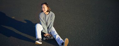 Mutlu Koreli genç kız kaykayının üstünde oturur, sörf tahtasında gezer, günlük kıyafetler giyer..