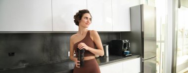Spor ve sağlıklı yaşam tarzı. Gülümseyen spor elbiseli bir kadının portresi, mutfak tezgahının yanında duruyor, su şişesinden içiyor, spor salonuna hazırlanıyor..