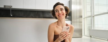 Aktif giyimli spor bir kadın görüntüsü, evde yoga minderinde oturuyor, akıllı telefonu tutuyor, egzersiz uygulaması kullanıyor, çevrimiçi egzersiz arıyor. Boşluğu kopyala
