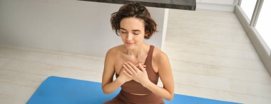 Düşünceli genç bir kadının portresi, çakrasını açıyor, evde yoga yapıyor, gözlerini kapıyor ve gülümsüyor, göğsünde huzuru hissediyor. Spor ve yaşam tarzı kavramı