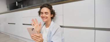 Mutlu kız yerde oturuyor ve akıllı telefon uygulamasında görüntülü sohbetler yapıyor, cep telefonuyla sohbete bağlanıyor, telefon kamerasına el sallıyor..