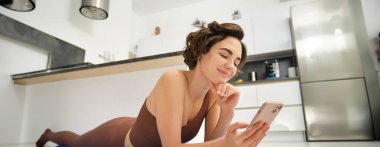 Çevrimiçi fitness ve refah. Yoga minderinde uzanan genç bir kadının portresi, akıllı telefon uygulamasına bakmak, egzersiz ipuçlarına bakmak, aktif kıyafet eğitimi almak..