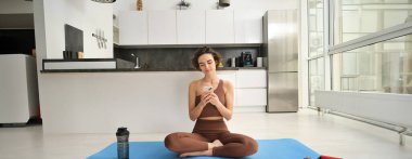 Spor uygulaması ve refah. Genç bir kadın, atlet akıllı telefonuyla evde oturuyor, yoga yapıyor, cep telefonunda egzersiz uygulaması kullanıyor, evde egzersiz yapıyor. Boşluğu kopyala
