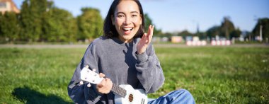 Müzik aleti olan sevimli Asyalı kız portresi. Şaşkın yüzlü, ukulele tutan ve parkta battaniyeye oturan genç bir kadın..