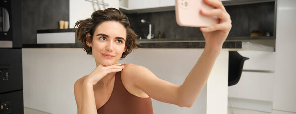 Спортивная молодая спортсменка, девушка делает селфи на смартфоне, тренируется из дома и делает фотографии, записывает видео по телефону во время фитнес-упражнений.