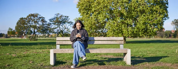 Young woman sitting alone on bench in park, using mobile phone, looking at screen, texting, sending messages in app chat.
