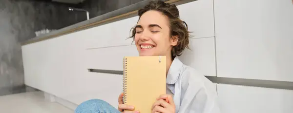 Portrait of happy, carefree woman, writes in her journal and laughs, holds her diary, reads something in notebook, sits on floor indoors. Lifestyle and leisure concept