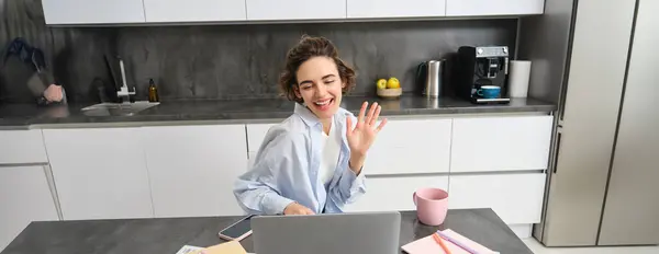 Happy young woman, tutor teacher students online. Girl connects to remote work meeting from her home laptop, says hello, waves hand at computer.