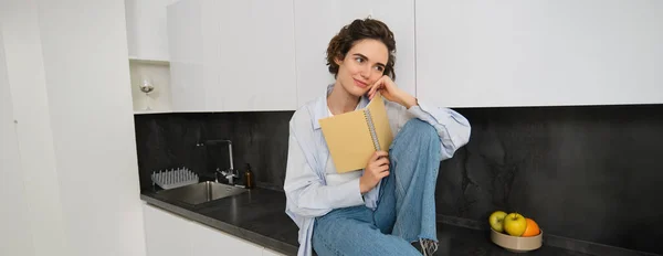 stock image Portrait of modern young woman reading in kitchen, sitting on counter and smiling, studying. Copy space