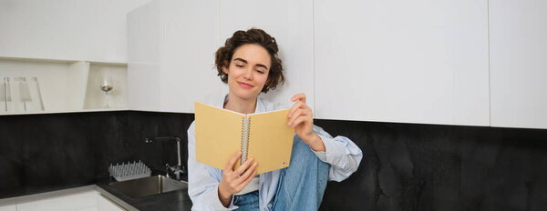 Концепция образа жизни и людей. Молодая женщина наслаждается выходными дома, читает заметки в своем дневнике, сидит на кухонном столе с ноутбуком и улыбается.