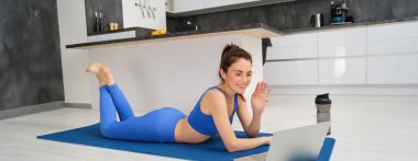 Mutlu genç bir kadının portresi, evde fitness yapıyor, bilgisayardaki görüntülü sohbete bağlanıyor, bilgisayara selam vermek için el sallıyor, yoga minderinde aktif giysiyle yatıyor..