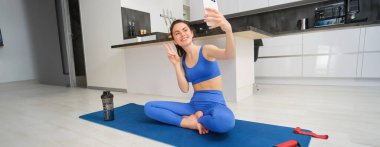 Genç gülümseyen bir kadın, fitess kız evden antrenman yapıyor, yoga minderinde akıllı telefonuyla oturuyor, selfie çekiyor, spor videolarını içeride kaydediyor..