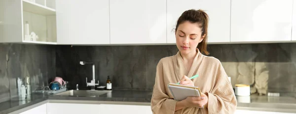 Portrait of woman writing grocery list for shopping, standing in the kitchen and making notes for recipe, cooking at home in bathrobe.