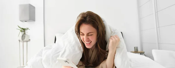 Woman with smartphone, feeling excitement, reading mobile phone and cheering, lying in bed under white blanket, using smartphone and looking happy in morning.