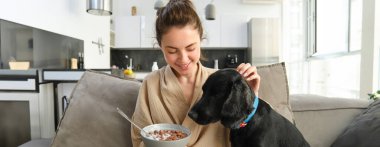 Bornozlu mutlu genç bir kadın, evde köpeğiyle vakit geçirmeyi seviyor, mısır gevreği yiyor, yavru köpekle kahvaltı ediyor, kanepede oturuyor..