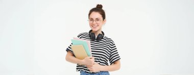 Eğitim konsepti. Gülümseyen esmer kız, günlük giysiler içinde öğrenci, kitaplarını tutuyor, ders çalışıyor, kulaklık takıyor, kendinden emin ve mutlu görünüyor, beyaz arka planda izole edilmiş..