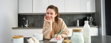 Çekici, genç, neşeli kız mutfakta yemek pişiriyor, para kazanıyor, yemek tarifi kitabını tutuyor, fikirleri var..