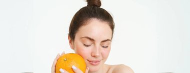 Esmer kadın modelin yakın portresi, turuncuyu yüzüne yakın tutuyor, beyaz arka planda duruyor..