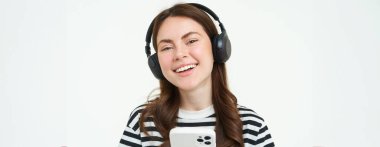 Güzel genç bir kadının portresi kablosuz kulaklıkla müzik dinliyor, cep telefonunu kullanıyor, gülümsüyor ve mutlu görünüyor, beyaz arka planda izole edilmiş..