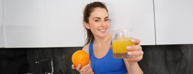 Gülümseyen, güzel genç bir kadının portresi, bir bardak taze meyve suyu sunuyor, elinde bir portakal tutuyor ve mutlu görünüyor, fitness eğitmeni sana içki veriyor..