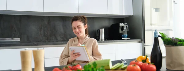 stock image Portrait of beautiful, smiling young woman making list of meals, writing down recipe, sitting in the kitchen with vegetables, doing house errands.