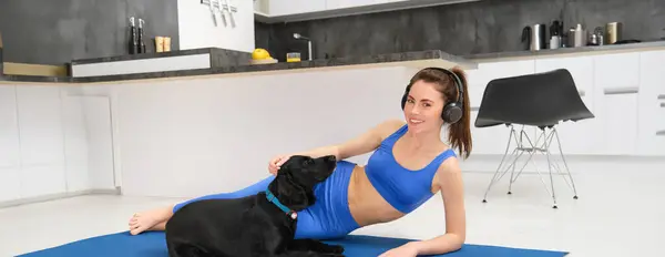 Portrait of beautiful young fitness woman, laying on yoga mat , workout at home, playing with her dog while doing sports. Lifestyle and wellbeing concept