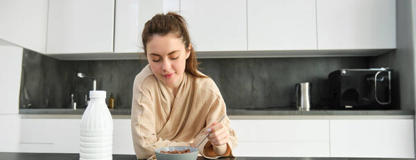 Portrait of beautiful woman at home, eating her breakfast, holding spoon, leaning on worktop and having cereals with milk.