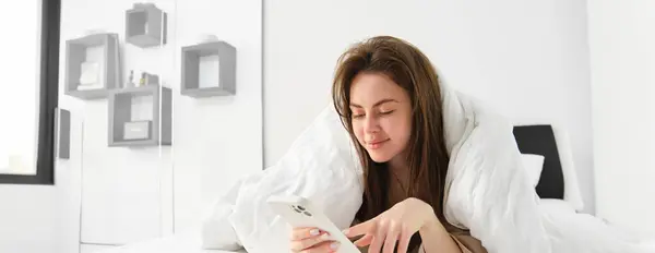 Woman in morning lying in bed covered in blanket, looking at smartphone, using mobile phone app.