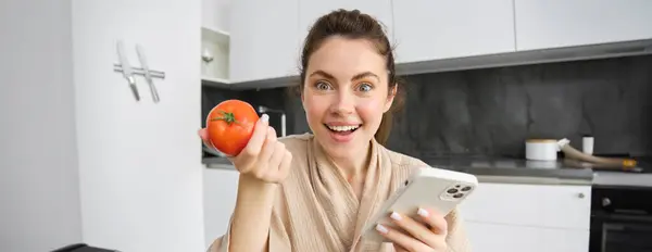 Close up portrait of beautiful young woman in the kitchen, holding tomato and smartphone, buys fresh vegetables online, orders groceries delivery on mobile phone application.
