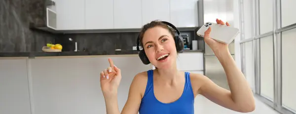 Close up portrait of happy fitness girl, listens music in wireless headphones, holds smartphone, dances and workout from home.