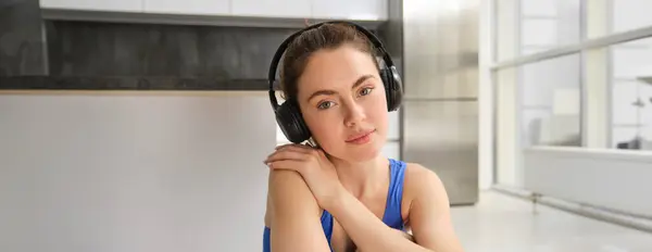 Close up portrait of brunette fitness girl, listens music in wireless headphones, smiles at camera, does workout alone at home, sits on floor and exercises.