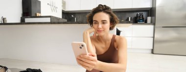 Teknoloji ve spor konsepti. Jimnastikçi kız akıllı telefonuna bakıyor, spor kıyafetleriyle evde oturuyor, cep telefonuyla egzersiz videoları izliyor..