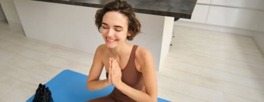 Genç fitness kızı portresi, evdeki lastik mindere oturmuş, namaste ellerle nilüfer çiçeği pozu veriyor, evde yoga yapıyor, meditasyon rehberini dinliyor. Spor ve farkındalık kavramı
