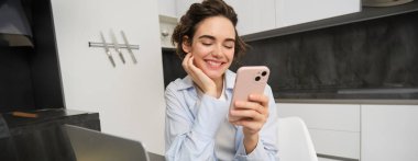 Evde akıllı telefonunu kullanan genç bir kadının görüntüsü. Kız cep telefonuyla mutfakta oturuyor ve gülümsüyor..