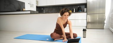 Gülümseyen genç bir kadının portresi, kauçuk yoga minderinde fitness eğitmeni, tabletten online eğitim sınıfına katılıyor, kablosuz kulaklıkla video dinliyor, evde yerde egzersiz yapıyor..