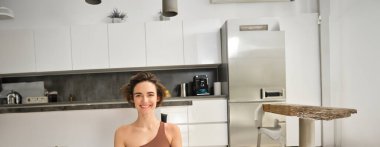 Sağlıklı, fit bir kızın dikey portresi, evde yoga yapmak, lotus pozu vermek ve gülümsemek, aydınlık odada egzersiz yapmak..