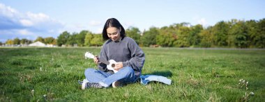 Çimenlerde ukulele çalan Asyalı kız şarkı söylüyor, parkta battaniyeye oturuyor, güneşli bir günde dışarıda dinleniyor..